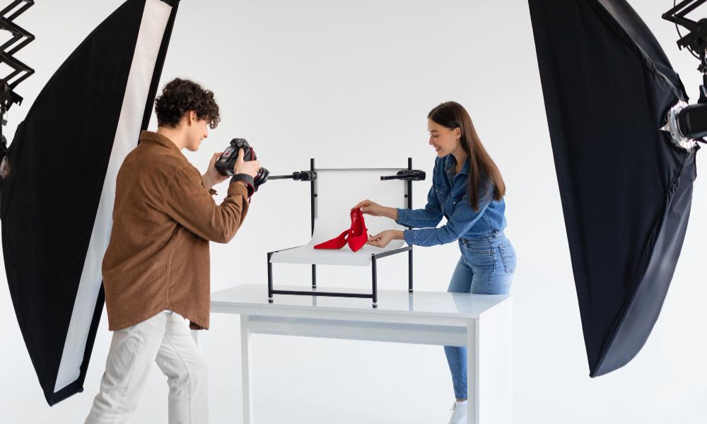 Young male photographer taking photo of stylish red shoes on white platform in photo studio with modern equipment