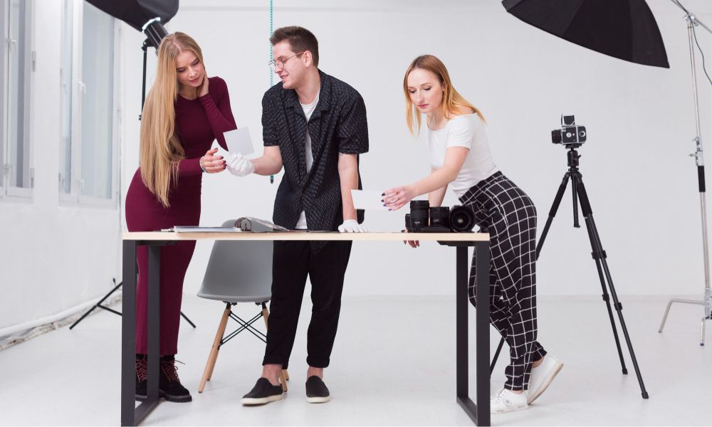 Three young adults are gathered around a table in a bright photo studio. A woman with long blonde hair in a maroon dress examines a print with a man in glasses and a black patterned shirt, who is wearing white gloves and also looking at prints. Another woman with blonde hair in a white t-shirt and plaid pants looks at a camera on the table next to a tripod