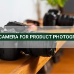 DSLR camera on a wooden surface with a blurred second camera in the background. Text: "BEST CAMERA FOR PRODUCT PHOTOGRAPHY"