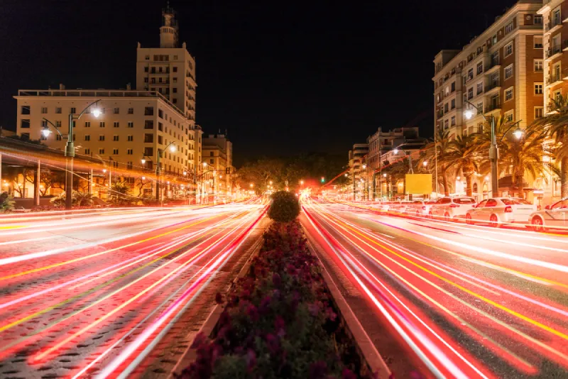 Long exposure photo of a busy city street at night, showing colorful light trails from moving vehicles with buildings illuminated on both sides