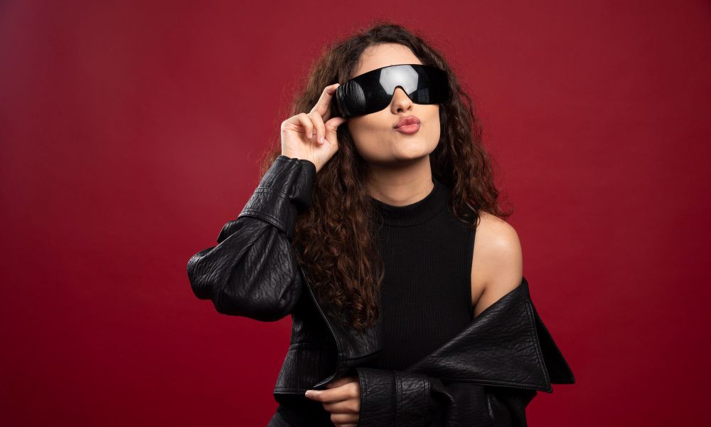 A fashionable woman with long, curly dark hair, wearing a black top and a black leather jacket slung over her shoulders