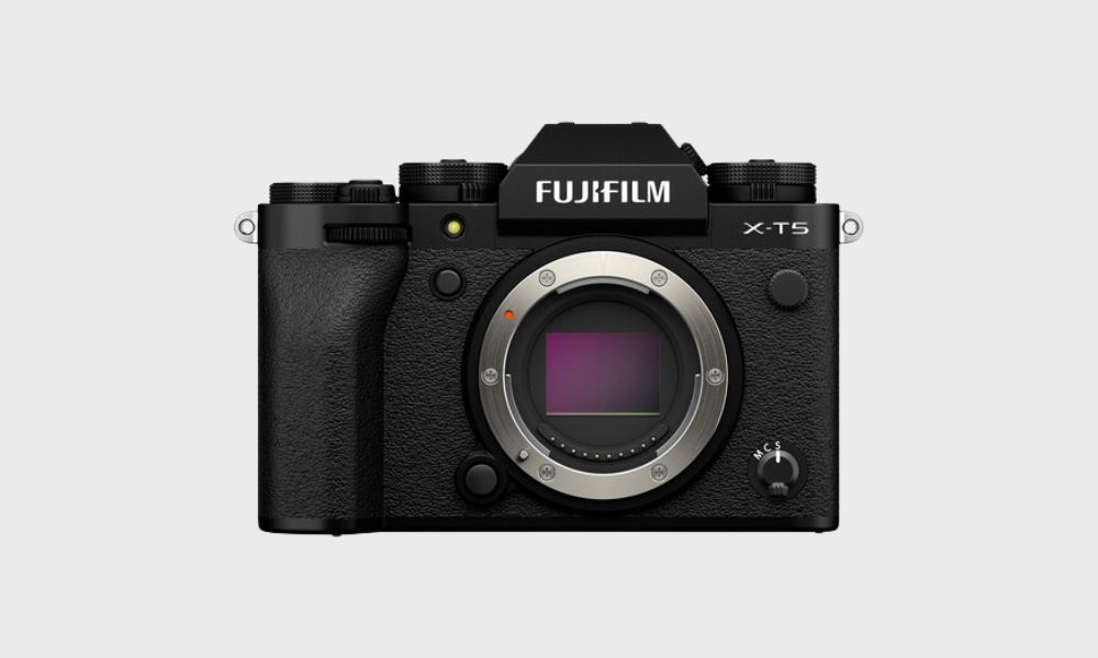 A black Fujifilm X-T5 mirrorless camera body, viewed from the front, with its lens mount cap visible and no lens attached, is set against a plain light gray background