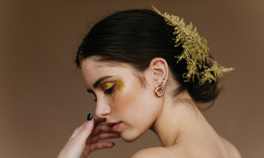 A glamour portrait of a woman with dark hair pulled back, adorned with golden glitter on her eyelids and cheekbones, and green foliage in her hair. She is wearing hoop earrings and has dark nail polish on one visible hand, which is touching her face.