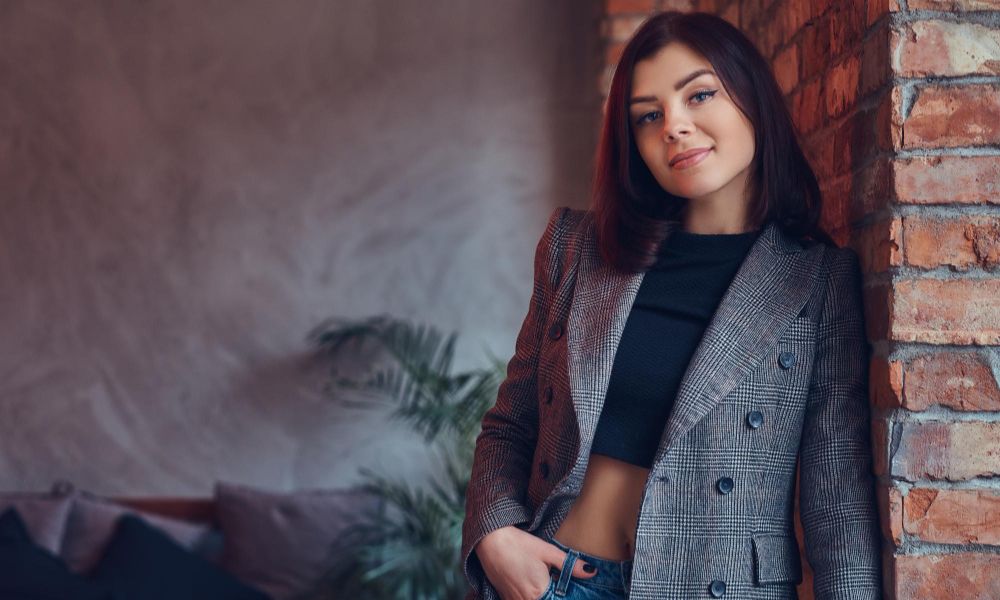 An indoor portrait of a slim young woman with dark hair, wearing a plaid double-breasted jacket over a black crop top and jeans, leaning casually against a brick wall and smiling towards the camera