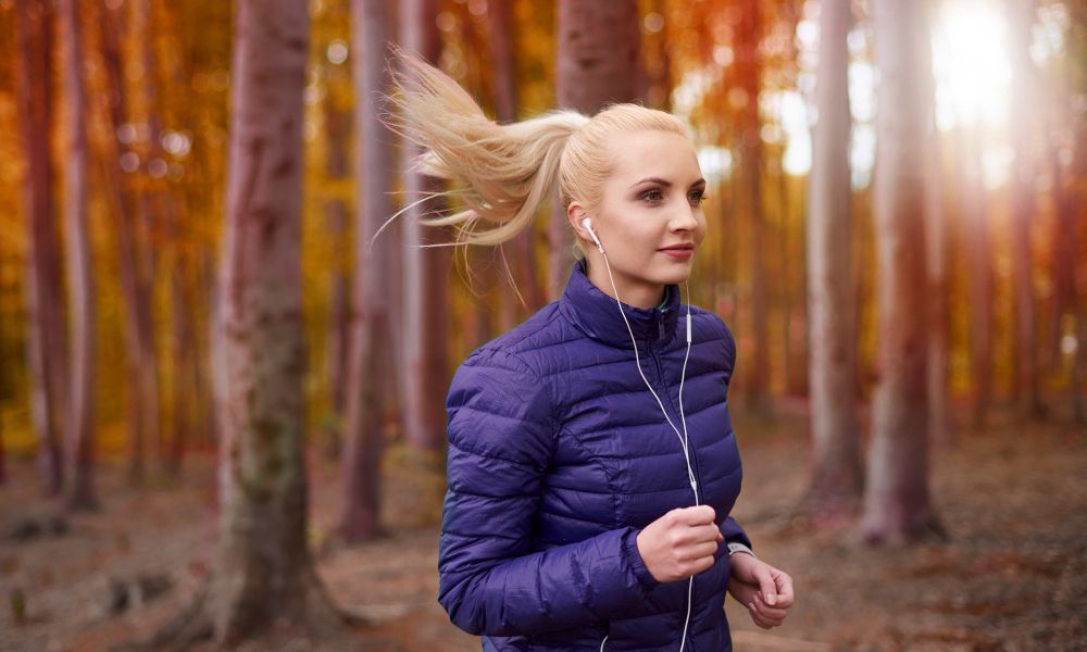 A blonde woman with her hair in a high ponytail, wearing a purple puffer jacket and white earphones, is jogging through an autumnal forest with warm-toned trees and sun flare in the background