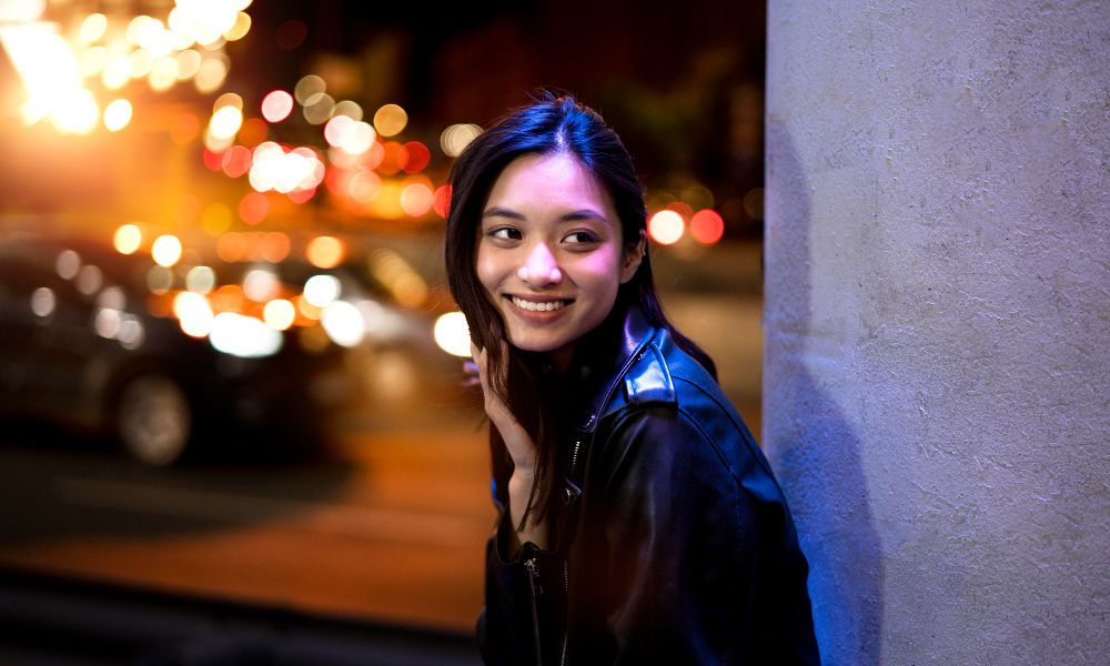 A vibrant night portrait of a young woman with long dark hair, wearing a black leather jacket, smiling and looking over her shoulder. She is illuminated by the purple glow from a pillar on the right, with blurred city lights and car headlights forming a warm, bokeh background
