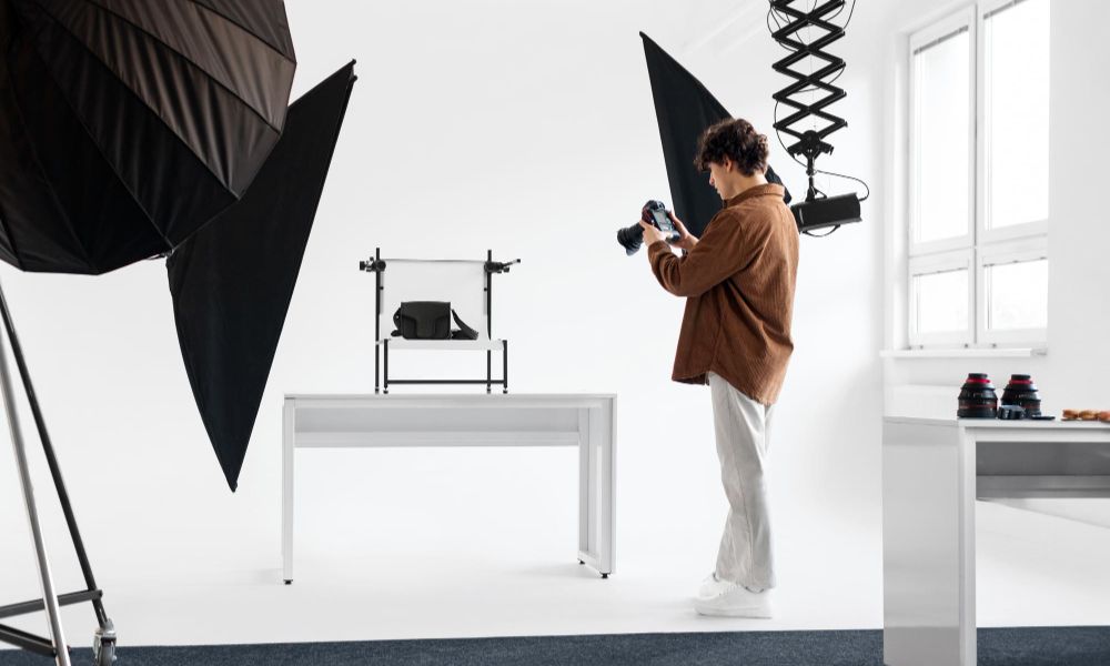 A photographer with curly hair, wearing a brown shirt and light pants, stands in a brightly lit studio, holding a camera and photographing a black bag set up on a white table with a white backdrop. Large black softboxes are positioned on either side of the setup