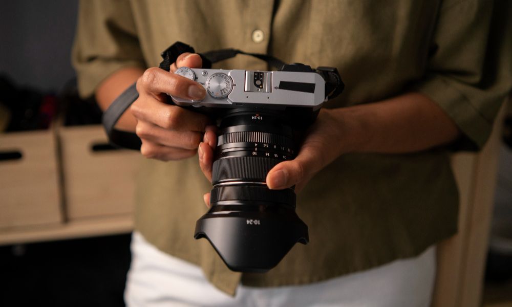 A person's hands are holding a silver and black camera with a large black lens, with their fingers positioned to adjust settings on the top dial. The person is wearing a muted olive-green shirt and white pants, with the background slightly blurred