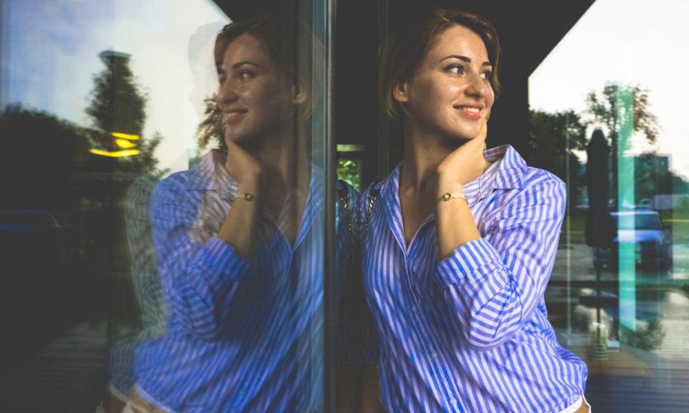 A smiling young woman in a blue and white striped shirt is captured in a half-body outdoor portrait, with her reflection visible on a large glass surface beside her. She is looking away and smiling