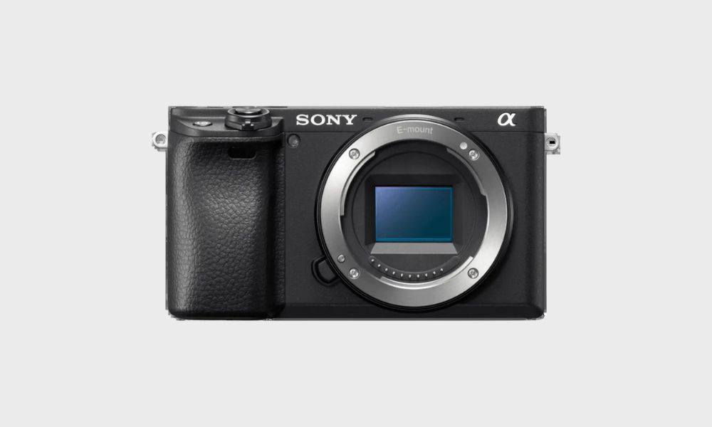 A black Sony A6400 mirrorless camera body, viewed from the front, with its E-mount lens opening and sensor visible, is set against a plain light gray background
