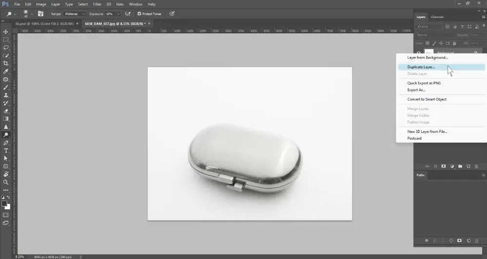 A screenshot of Photoshop shows a metallic, oval-shaped object, possibly a pill case, on a white background. A contextual menu is open, with the "Duplicate Layer..." option highlighted. The layers panel is visible on the right