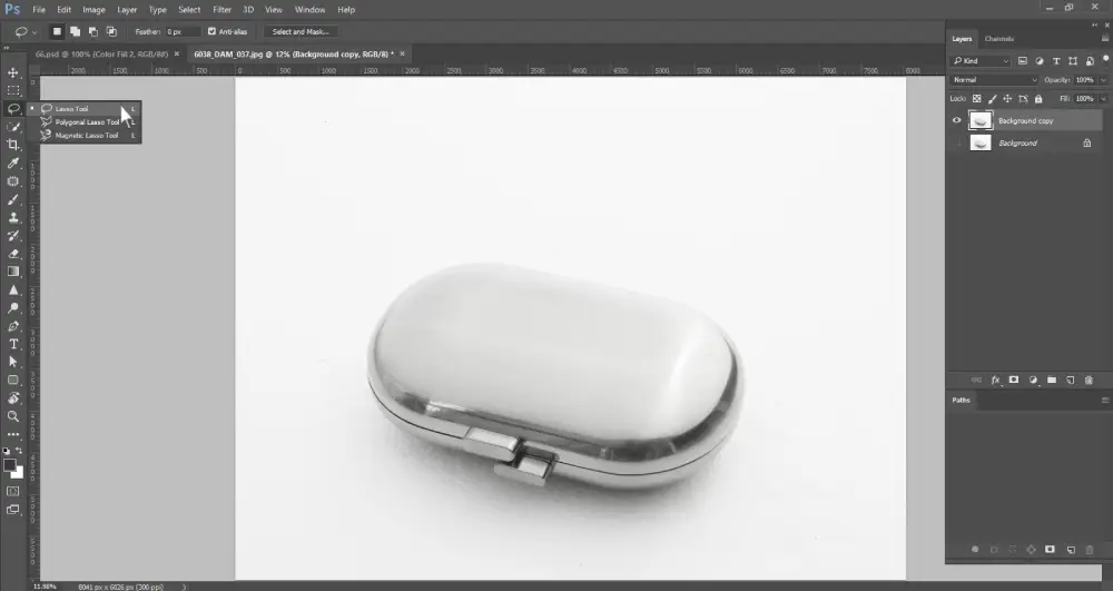A Photoshop screenshot shows a silver, oval-shaped object, possibly a pill case, against a white background. A dropdown menu on the left toolbar displays "Lasso Tool," "Polygonal Lasso Tool," and "Magnetic Lasso Tool," with "Lasso Tool" currently selected. The layers panel on the right shows "Background copy" and "Background" layers