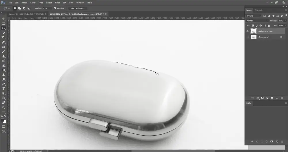 A screenshot from Photoshop shows a metallic, oval-shaped object, possibly a pill case, on a white background. A jagged, dotted line (characteristic of the Lasso Tool) is being drawn over the top surface of the object, indicating an active selection process. The layers panel on the right shows "Background copy" and "Background" layers