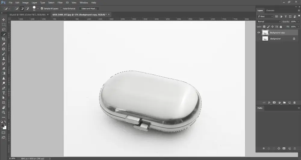 A Photoshop screenshot displays a metallic, oval-shaped object, possibly a pill case, on a white background. A dotted selection outline (marching ants) perfectly traces the perimeter of the object, indicating it has been fully selected using a lasso tool. The layers panel on the right shows "Background copy" and "Background" layers