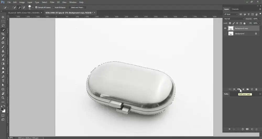 A Photoshop screenshot displays a metallic, oval-shaped object, possibly a pill case, on a white background, enclosed by a dotted selection outline. The cursor hovers over the "Add layer mask" icon at the bottom of the layers panel, which is highlighted. The layers panel shows "Background copy" and "Background" layers