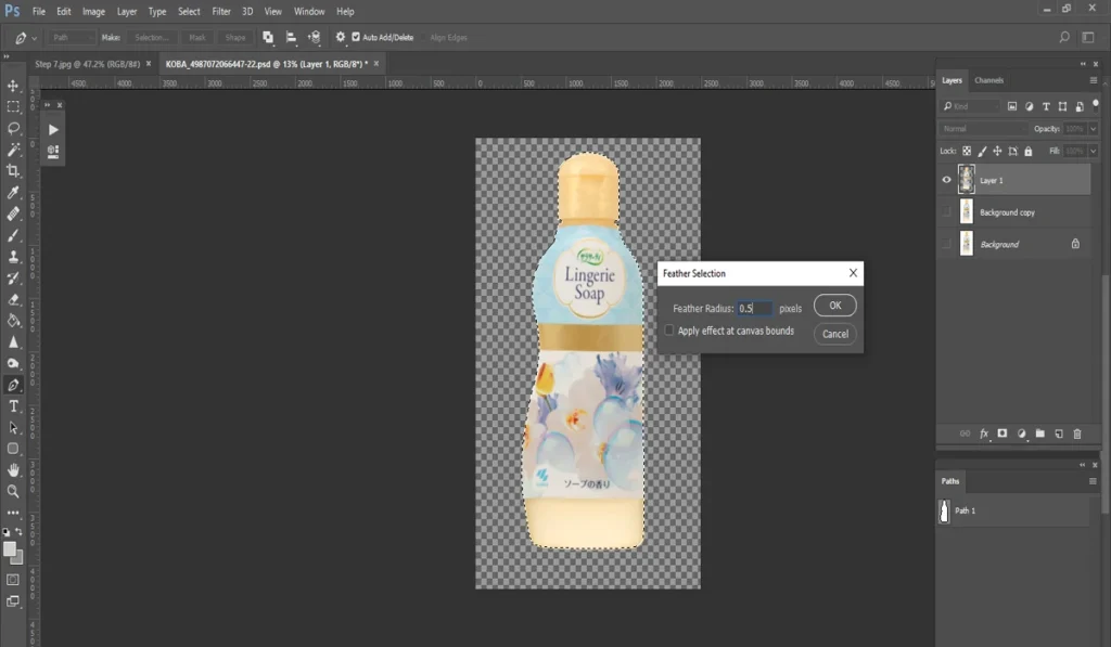 A screenshot of Adobe Photoshop shows a bottle of "Lingerie Soap" with a floral design on a checkered background, and a "Feather Selection" dialog box open, set to a radius of 0.5 pixels