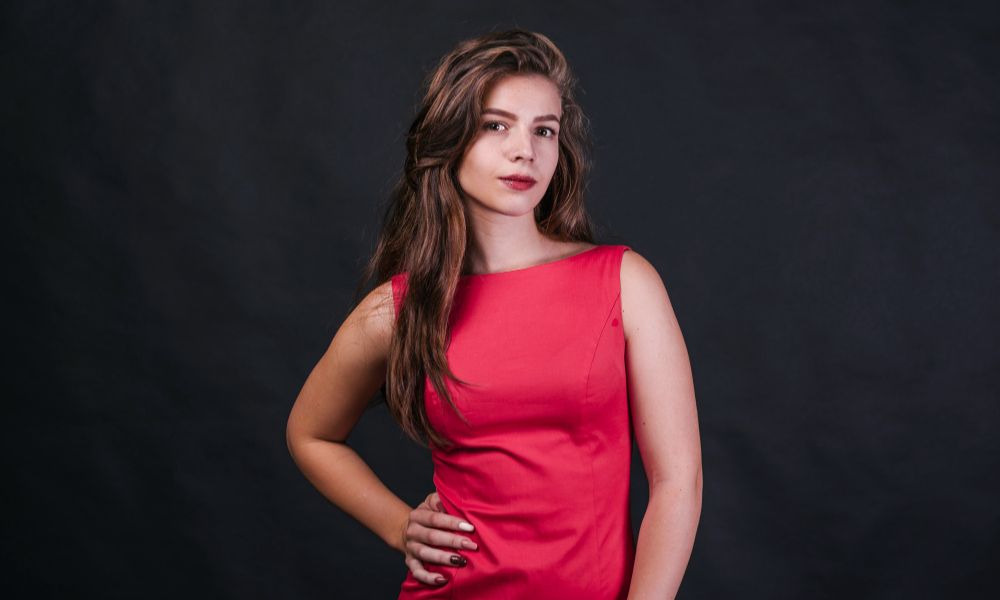 A studio portrait of a young woman with long, wavy brown hair, wearing a sleeveless pink dress, standing with one hand on her hip and looking directly at the viewer with a subtle smile, against a dark grey background