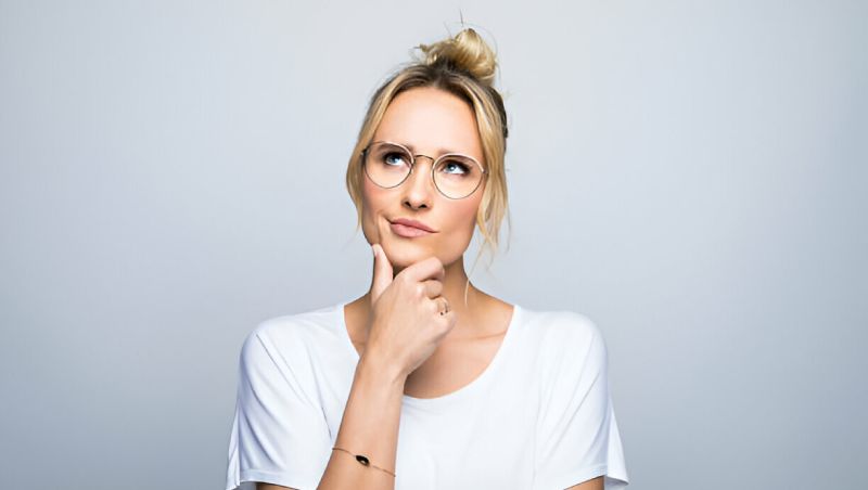 Thoughtful young woman with blonde hair in a bun, wearing round glasses and a white t-shirt, holding her chin while looking up to the side against a plain light gray background