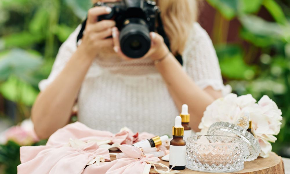 A female photographer in a white top with long blonde hair, holding a black DSLR camera up to her face, takes a picture of various cosmetic products arranged on a wooden table