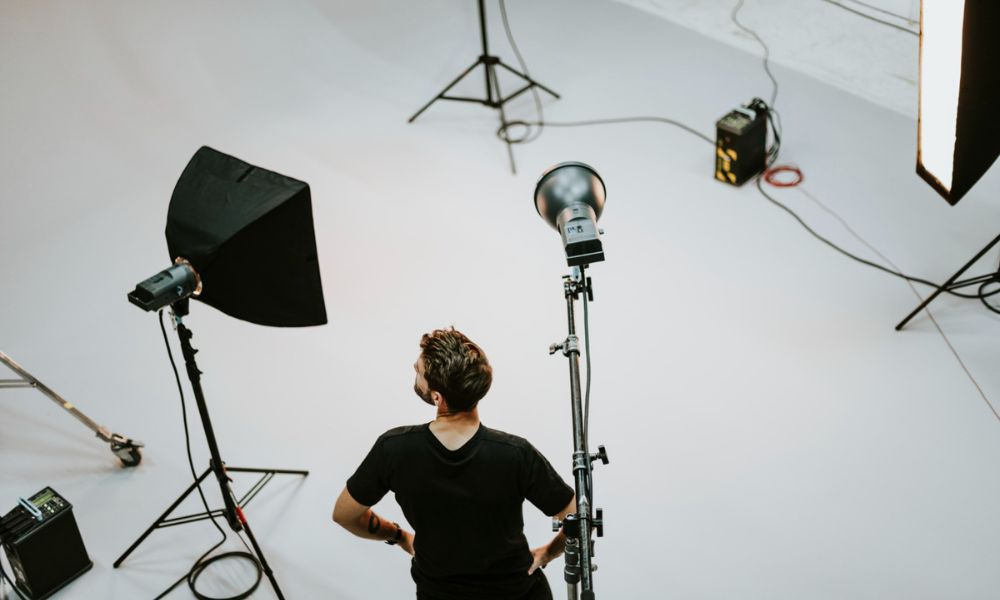 An overhead shot shows a man standing in a photography studio with his hands on his hips, facing away from the camera, surrounded by various studio lighting equipment and light stands on a white floor