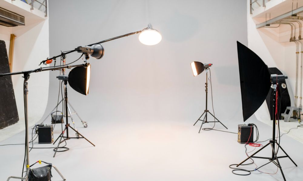 A professional photography studio features a seamless white backdrop and floor, with various studio lights and stands positioned around the space, including a large overhead light and several softboxes and reflectors