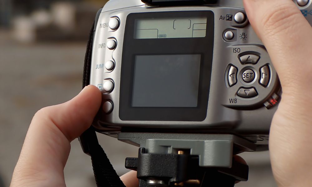 A close-up view shows a person's hands adjusting the settings on the back of a silver DSLR camera, with various buttons and a small display screen visible