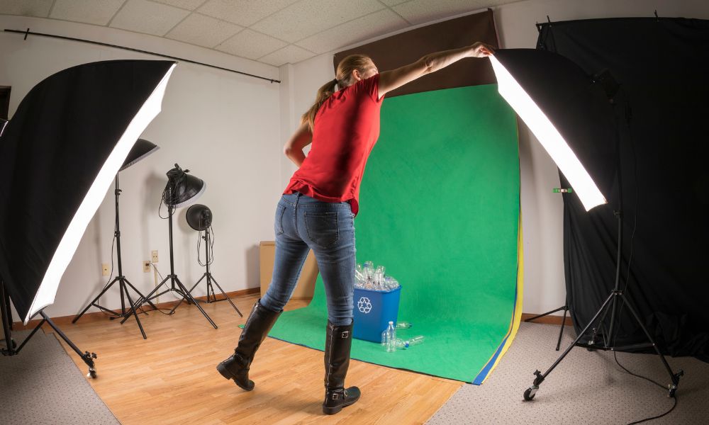 A woman in a red t-shirt and blue jeans adjusts a green backdrop in a photo studio, where a blue recycling bin filled with plastic bottles sits, flanked by two large studio softbox lights