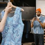 A person holds up a blue patterned top on a hanger while another person in an orange beanie photographs it with a smartphone in a clothing store