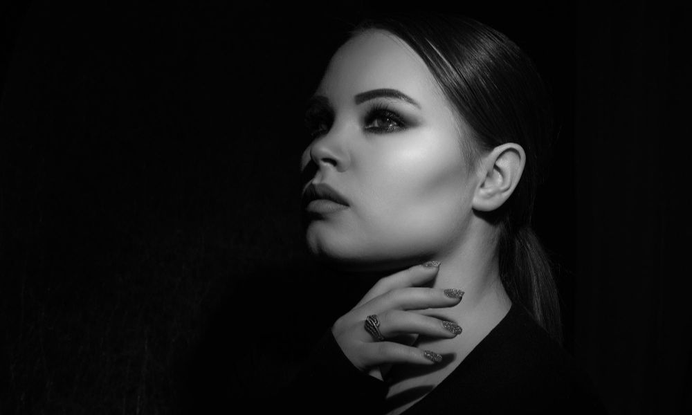 A high-contrast, black and white portrait of a young woman with a sleek ponytail, dramatic eye makeup, and a ring, with one hand gently touching her neck, looking upwards and to her left against a dark background
