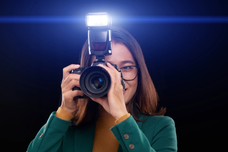 Woman holding a DSLR camera with an external flash firing, wearing glasses and a green blazer against a dark background