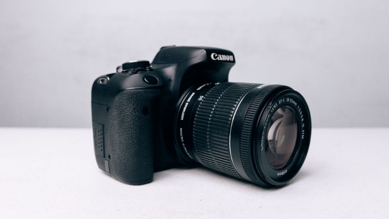 A black Canon DSLR camera with an attached zoom lens is placed on a light-colored surface with a blurred white background