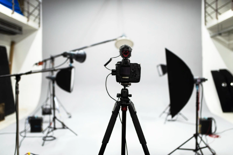 Digital camera mounted on a tripod in a professional photography studio setup, surrounded by softbox lights, reflectors, and a boom microphone, all against a white seamless background