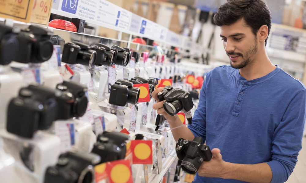 A young man with dark hair and a beard, wearing a blue long-sleeved shirt, stands in an electronics store, examining two DSLR cameras while surrounded by shelves displaying many more cameras