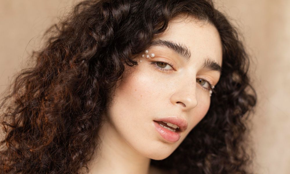 A close-up portrait of a woman with dark, curly hair, fair skin, and natural makeup. She has small pearls delicately placed near the outer corners of both eyes, and her lips are slightly parted