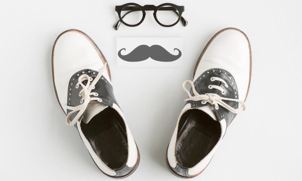 A pair of black and white saddle shoes are displayed on a white background, with black-framed glasses above them and a stylized black mustache in between