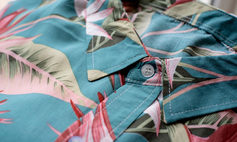 A close-up of a blue and pink Hawaiian-style shirt with a tropical leaf pattern and a visible white button on the collar