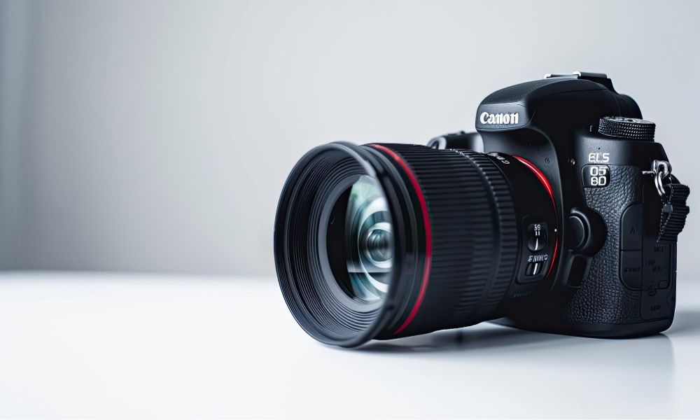 A close-up side view of a black Canon DSLR camera with a lens, featuring red accents and clear lens reflections, resting on a white surface against a light grey background