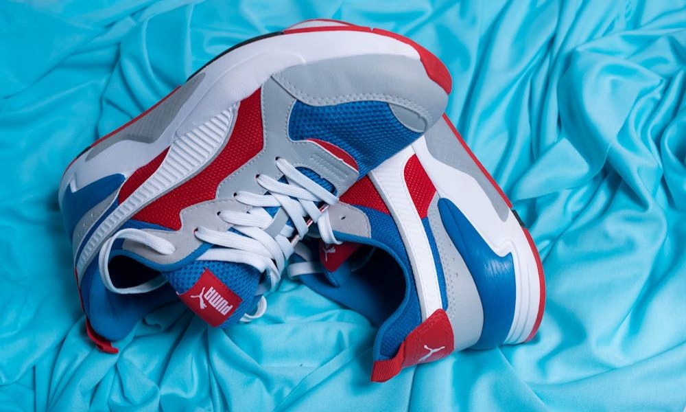 A pair of red, white, and blue Puma sneakers with white laces are placed on a crumpled blue fabric surface