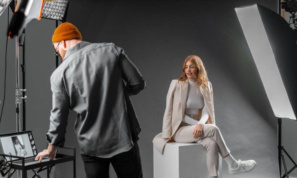 A professional studio setup with a male photographer in an orange beanie and glasses looking at a laptop, while a female model in a cream suit and white sneakers sits on a white cube. Studio lights are prominent on either side