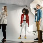A clothing photoshoot in progress: a stylist, a model in a red jacket, and a photographer with a camera and flash umbrella in a studio with a white backdrop