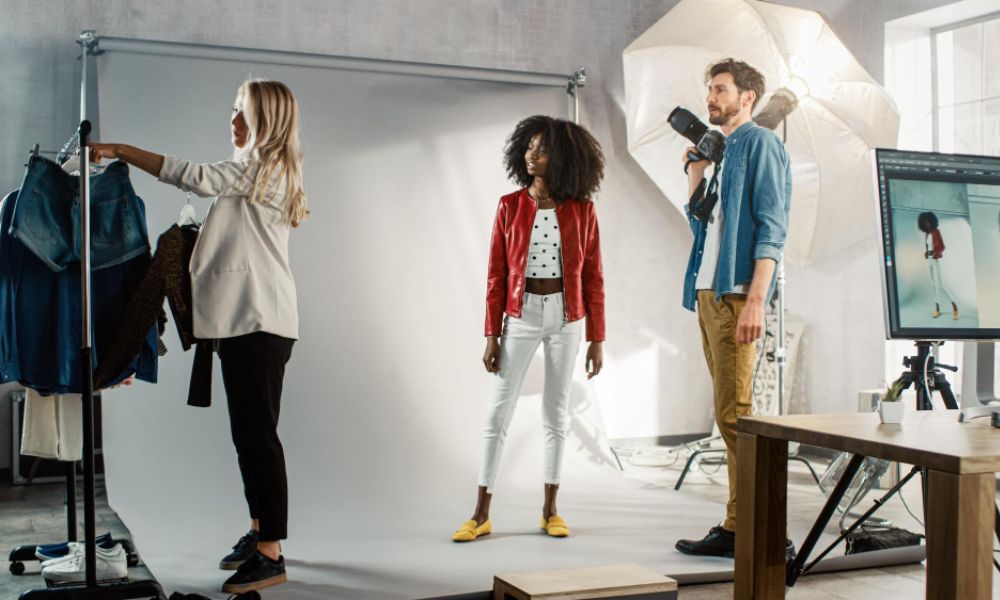 A clothing photoshoot in progress: a stylist, a model in a red jacket, and a photographer with a camera and flash umbrella in a studio with a white backdrop