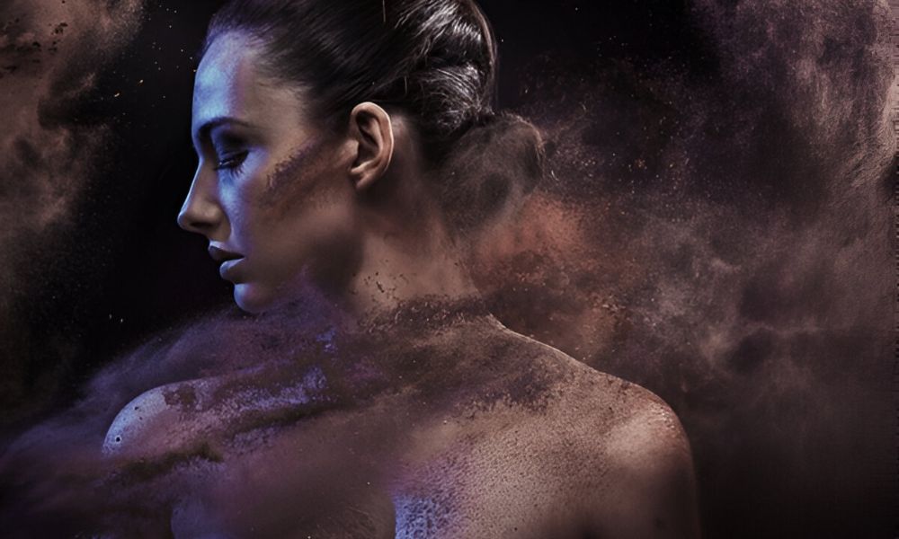 A conceptual photograph of a woman's profile, with her hair pulled back, as if she is partially dissolving into a cloud of purple and brown dust and particles against a dark background
