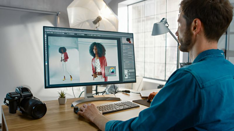 Man sitting at a desk using a computer to edit a fashion photo of a woman in a red jacket and white pants, with studio lights and a camera visible in the background