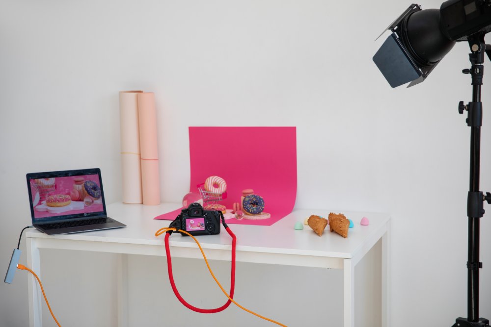 DIY home studio setup with a camera, laptop, lighting, and pink backdrop for photographing donuts and pastries on a white table