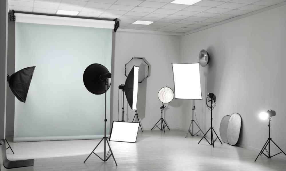 A professional photography studio is shown with a light blue seamless backdrop on the left and various lighting equipment, including softboxes, umbrellas, and reflectors, scattered across the grey floor