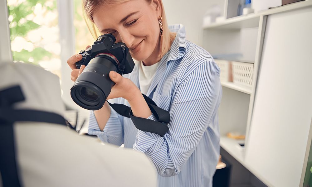 A blonde female photographer wearing a striped long-sleeved shirt is smiling and looking through the viewfinder of her camera, capturing a close-up shot of a white garment in a bright room with shelves in the background