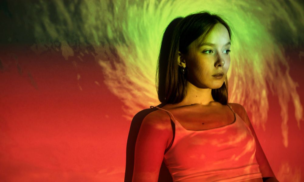 A fine art studio portrait of a woman with short dark hair, wearing a light-colored top, looking thoughtfully to her right. Her face and the background are illuminated by a striking green and red light projection, creating a swirling, abstract pattern