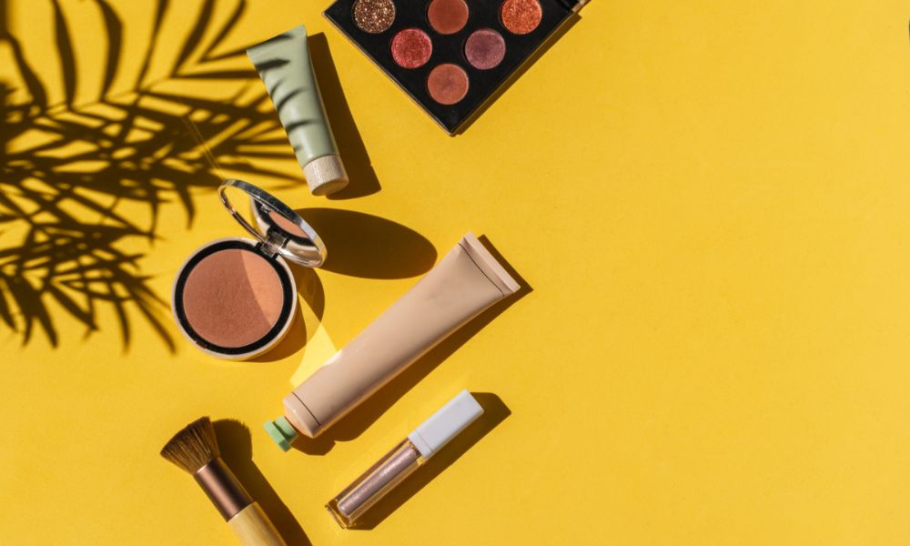 A flat lay shows various makeup products including an eyeshadow palette, two tubes of cream, a compact powder with a mirror, a lip gloss, and two makeup brushes, all arranged on a vibrant yellow background with palm leaf shadows
