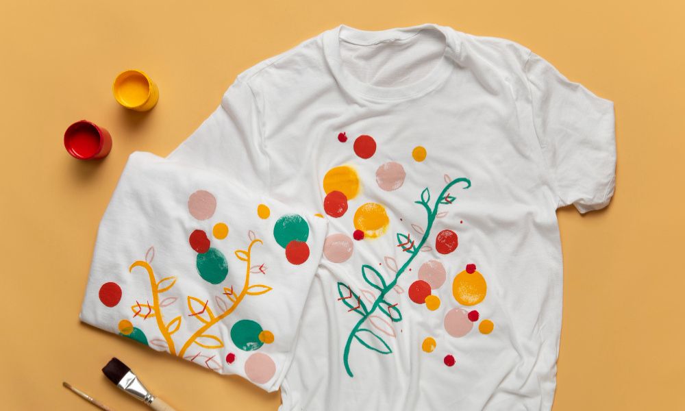 A flat lay shows two white t-shirts with colorful hand-painted designs of polka dots and branches. Next to them are small jars of red and yellow paint and a paintbrush, all on a light orange background