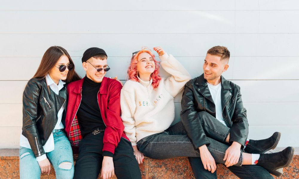 A diverse group of four young adults sitting casually on a ledge against a white wall
